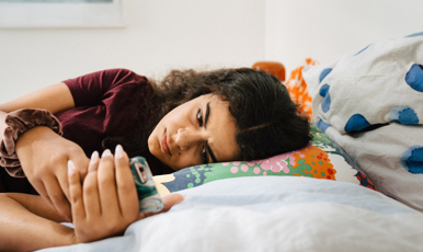 Tjej ligger på sin säng och tittar på telefonen med orolig blick.